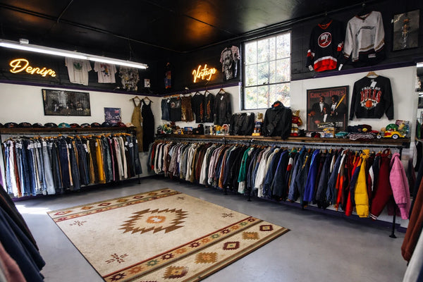Vintage clothing store with racks of clothes and neon signs on a patterned rug.